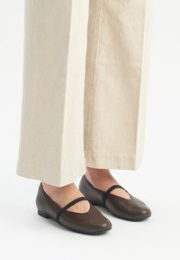 Ballerine in pelle marrone con punta arrotondata e cinturino elastico. Abbinati a pantaloni a gamba larga beige chiaro, che mostrano un design semplice.