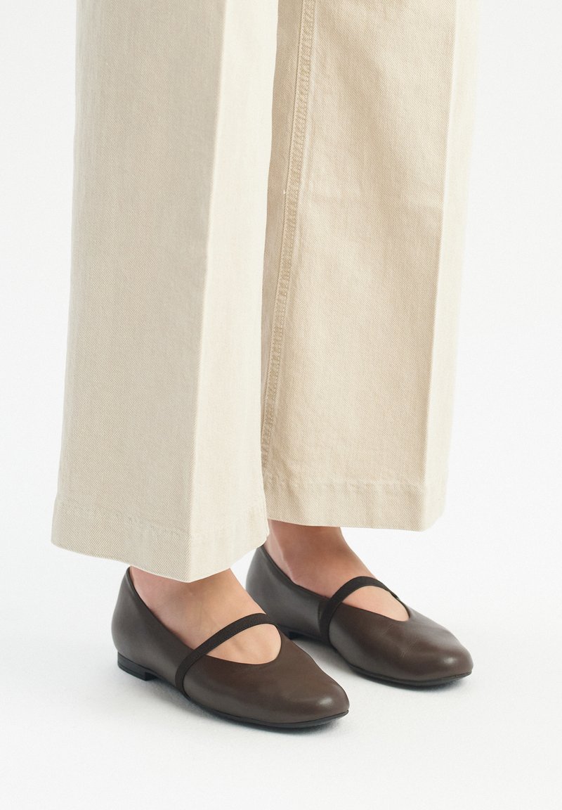 Ballerine in pelle marrone con punta arrotondata e cinturino elastico. Abbinati a pantaloni a gamba larga beige chiaro, che mostrano un design semplice.