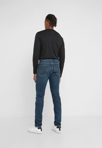 Camisa negra de manga larga con una costura trasera, combinada con jeans ajustados azules y zapatillas blancas con detalles en negro. Diseño minimalista, textura suave.