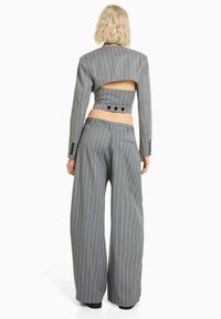 Vrouw met kort blond haar die een grijze streepjessuit met wijde pijpen draagt, compleet met een cropped jasje met een uitsparing aan de achterkant en knoopdetails.