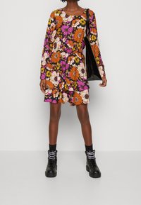 Blumenkleid mit langen Ärmeln und rundem Ausschnitt, in den Farben Orange, Pink und Weiß auf schwarzem Hintergrund; kombiniert mit schwarzen Stiefeln.