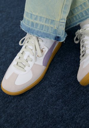 Baskets Puma blanches et beiges avec semelles en gomme portées avec un jean bleu délavé à ourlet effiloché sur un sol moquette foncé.