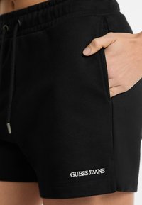 Juodi medvilniniai šortai su elastingu juosmeniu ir šoninėmis kišenėmis. Išsiskirianti balta "GUESS JEANS" siuvinėta etiketė apatinėje hem.