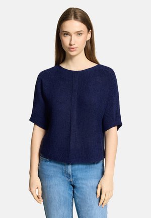 Jeune femme aux cheveux châtains lisses portant un pull en maille bleu marine à manches courtes et un jean bleu clair, debout devant un fond blanc.