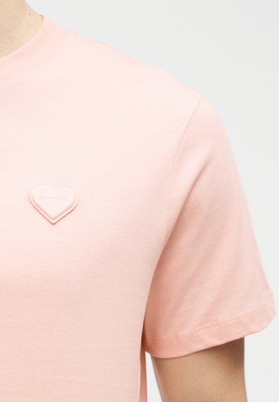 Κοντινό πλάνο ενός ατόμου που φοράει ανοιχτό ροζ κοντομάνικο πουκάμισο με ένα μικρό μπαλώμα σε σχήμα καρδιάς στο στήθος.
