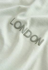 Světlá šedá látka s měkkou texturou, na které je vyšitý nápis "LONDON" ve světle tmavší šedé barvě. Design je jednoduchý a moderní.