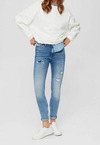 Jeans ajustados de un azul claro con detalles deshilachados, combinados con un sudadera blanca de gran tamaño y zapatillas blancas, mostrando un look casual.