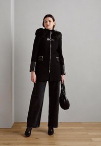 Cappotto nero con colletto in pelliccia sintetica, zip argentata e tasche laterali. Abbinato a pantaloni larghi scuri e una borsa nera. Stivali con tacco a spillo.