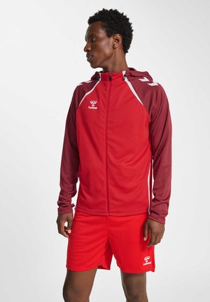 Jeune homme en veste de sport rouge et short, debout et regardant sur le côté, sur un fond blanc uni.