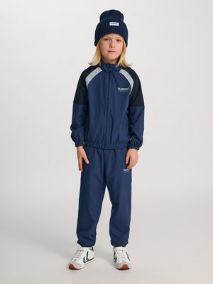 Barn står iført navyblå træningsdragt med sorte og grå accenter, navy hue og hvide sneakers med grønne detaljer på ensfarvet baggrund.