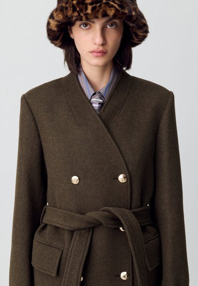 Cappotto doppiopetto verde oliva con bottoni dorati, vita aderente, e dettagli delle tasche, abbinato a una cravatta a righe e un cappello a stampa leopardata marrone.