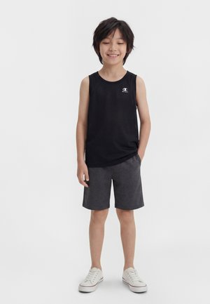 Nasmejan otrok stoji z eno roko v žepu, oblečen v črn brezrokavnik, temno sive kratke hlače in bele superge na enobarvnem belem ozadju.