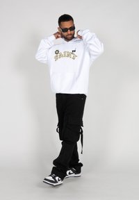 Witte hoodie met geborduurd "SAINT" in goud en zwart, gecombineerd met zwarte cargo pants met verstelbare banden en zakken, en zwart-witte sneakers.