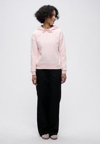 Világos rózsaszín kapucnis pulóver húzózsineggel, laza szabású, hosszú ujjú, laza fekete nadrággal és világos színű lábbelivel párosítva.