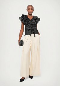 Schwarzes, gerafftes Oberteil mit Taillenschnürung, kombiniert mit weit geschnittenen, cremefarbenen Hosen. Das Outfit hat eine glatte Textur und wird durch schwarze High Heels ergänzt.