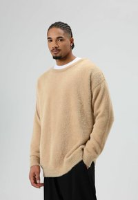 Beige oversized trui van zacht, gestructureerd materiaal, met een brede halslijn en geribbelde manchetten, gecombineerd met zwarte wijde broek.