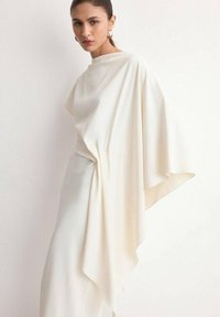 Woman wearing an elegant, flowing off-white dress with asymmetrical draped sleeves, standing against a plain white wall.