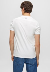 Camiseta blanca de manga corta en suave algodón con cuello redondo. Presenta un pequeño logo negro "HUGO" en la parte trasera del cuello. Combinada con unos vaqueros azules.