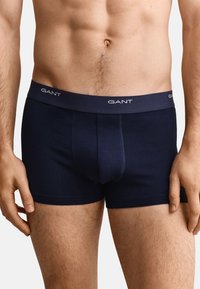 Buste masculin portant un boxer GANT bleu marine avec logo sur la ceinture, debout, les mains détendues le long du corps sur fond blanc.