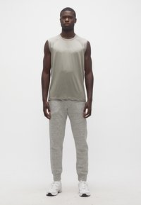 Άνδρας που στέκεται μπροστά, φορώντας αμάνικο γκρι πουκάμισο, ανοιχτό γκρι φόρμα και λευκά αθλητικά παπούτσια σε απλό λευκό φόντο.