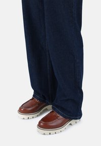 Zapatos de cuero marrón con una suela ancha blanca, combinados con pantalones de mezclilla de pierna ancha azul oscuro. Se puede ver el detalle de la costura en los zapatos.