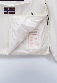 Hvit jakke med mesh-innlegg, med synlig vaskeanvisning og logo-patch. Inkluderer glidelåser og merkelapper for vannresistens.