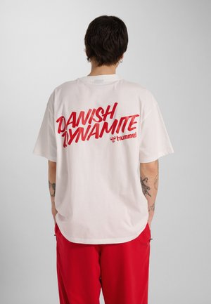 Person med kort mørkt hår iført hvid T-shirt med rød "Danish Dynamite" og "hummel" logo, parret med røde bukser, vender ryggen til.