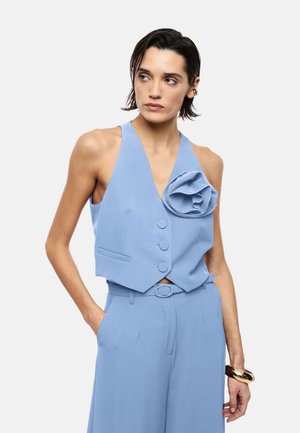 Donna con capelli corti e scuri che indossa un gilet azzurro chiaro senza maniche con un grande dettaglio floreale e pantaloni a vita alta abbinati, braccialetto d'oro al polso.