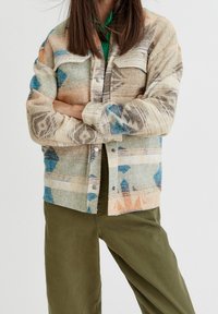 Veste tissée multicolore en beige, bleu et orange avec un motif géométrique ; elle comporte un devant à boutons-pression et deux poches poitrine.