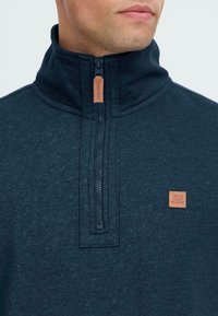 Moški nosi temno modro pulover s četrtno zadrgo, ki ima usnjeno zadrgo in majhen usnjen logotip na prsih.