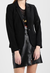 Femme portant un blazer noir sur un top graphique en maille et une mini-jupe en cuir noir avec une fermeture éclair frontale, se tenant devant un fond uni.