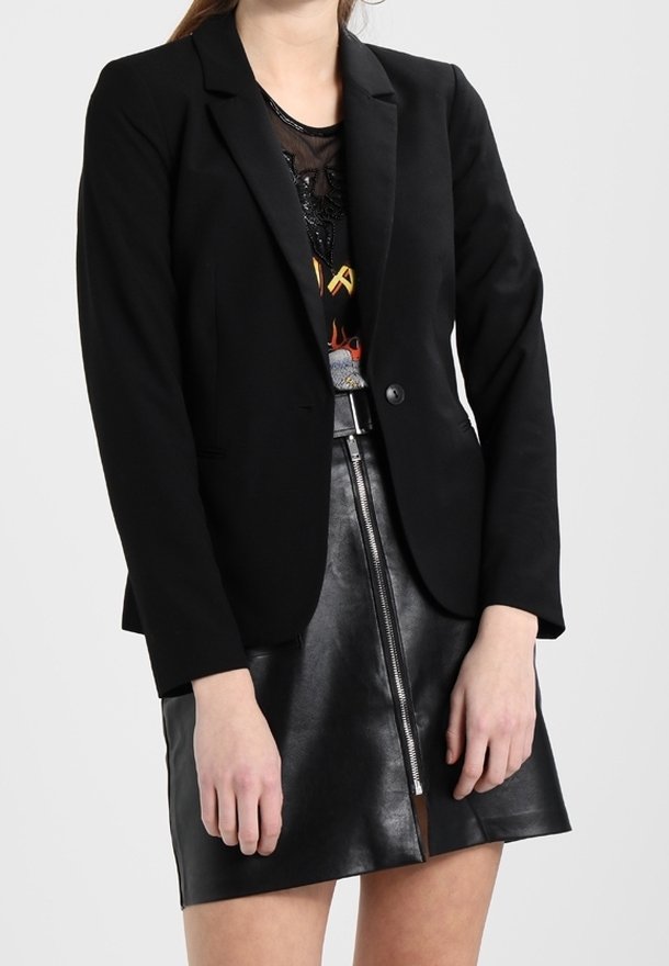 Femme portant un blazer noir sur un top graphique en maille et une mini-jupe en cuir noir avec une fermeture éclair frontale, se tenant devant un fond uni.