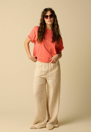 Femme aux longs cheveux ondulés portant des lunettes de soleil rondes, un t-shirt corail, un pantalon beige ample et des chaussures tissées beiges sans lacets, debout devant un fond uni.