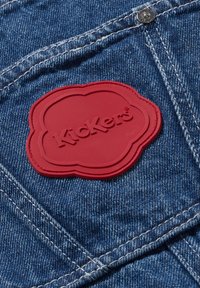 Tessuto denim con lavaggio blu caratterizzato da un patch in gomma rossa con il logo Kickers. Il design è arricchito da cuciture visibili e dettagli in metallo.