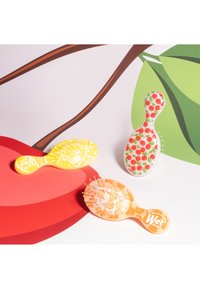 Trois petits brosses à cheveux Wet Brush colorées avec des motifs de fruits : citrons, cerises et oranges, sur un fond avec des formes rouges et vertes.