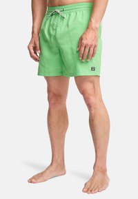 Άνδρας που φοράει φωτεινά πράσινα μαγιό σορτς με κορδόνι στη μέση, όρθιος ξυπόλητος μπροστά σε απλό φόντο.