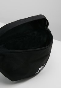 Trousse de toilette en tissu noir avec fermeture éclair. Texture douce, forme arrondie, et logo blanc de la marque à l'avant. L'intérieur est doublé et sombre.