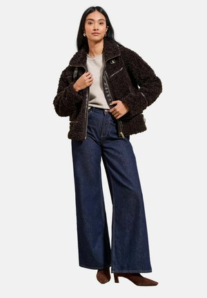 Kobieta w ciemnobrązowej, teksturowanej kurtce na beżowej bluzce, szerokich niebieskich dżinsach i brązowych butach na obcasie stojąca na białym tle.