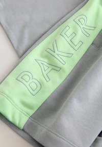 Sudadera gris con una amplia banda de color verde claro que presenta el texto en relieve "BAKER". Textura suave, diseño atlético, bloque de color contrastante.