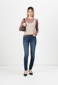 Beige mouwloos topje over een bruin shirt met lange mouwen, blauwe skinny jeans en zwarte ballerina's, met een kleine zwarte handtas in de rechterhand.
