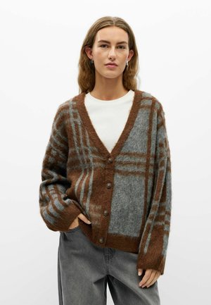 Jeune personne portant un cardigan boutonné à carreaux marron et gris par-dessus une chemise blanche et un pantalon gris ample, debout avec une main dans la poche.