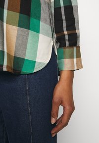 Camisa de franela a cuadros verde y negra con dobladillo curvado y puños abotonados, combinada con jeans de mezclilla oscura. Mano parcialmente visible.