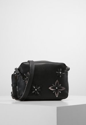 Sort læder crossbody taske med justerbar rem, blomsterformede metaldekorationer og diskret prægede brandlogo på en hvid piedestal.