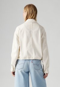 Kort cremefärgad denimjacka med krage, synlig sömnad och elastisk nederkant. I kombination med ljusblå vida jeans.