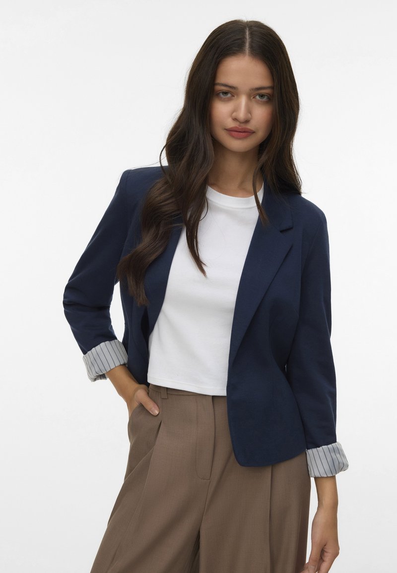 Jūras spēku ziepju jaka ar strukturētu piegriezumu, vienas pogas aizdari un svītrainām aprocēm, uzvilkta virs baltā T-kreklu un brūnām biksēm.