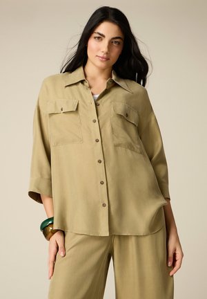 Donna dai lunghi capelli scuri con una camicia beige larga con bottoni e pantaloni larghi abbinati, in posa su uno sfondo semplice.