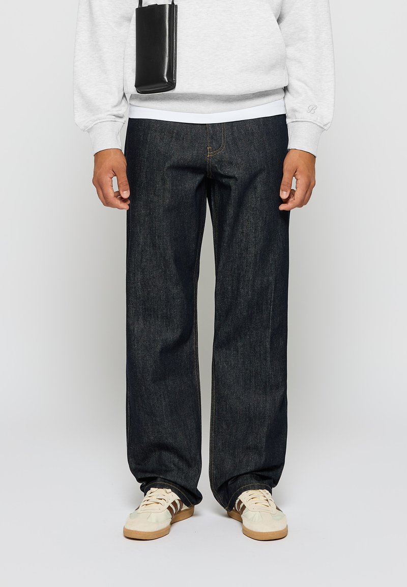 Mørkeblå denimjeans med rett benpassform, med subtile sømmer og et middels tungt stoff. Kombinert med kremfargede joggesko og en grå genser.