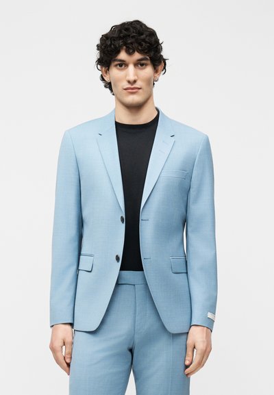 Νεαρός άνδρας με σγουρά μαλλιά ντυμένος με ανοιχτό μπλε κοστούμι και μαύρο πουκάμισο, στέκεται μπροστά σε μια απλή λευκή επιφάνεια.