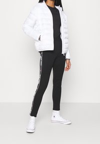 Giacca bianca imbottita, t-shirt nera, leggings neri con strisce del logo sui lati e sneakers alte bianche. Tessuto liscio e design aderente.