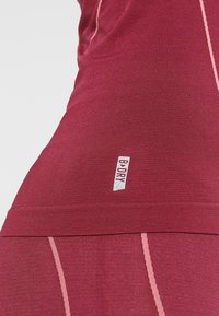 Gros plan sur un haut de sport bordeaux avec des coutures rose clair et un petit logo "B·DRY" sur le côté inférieur.